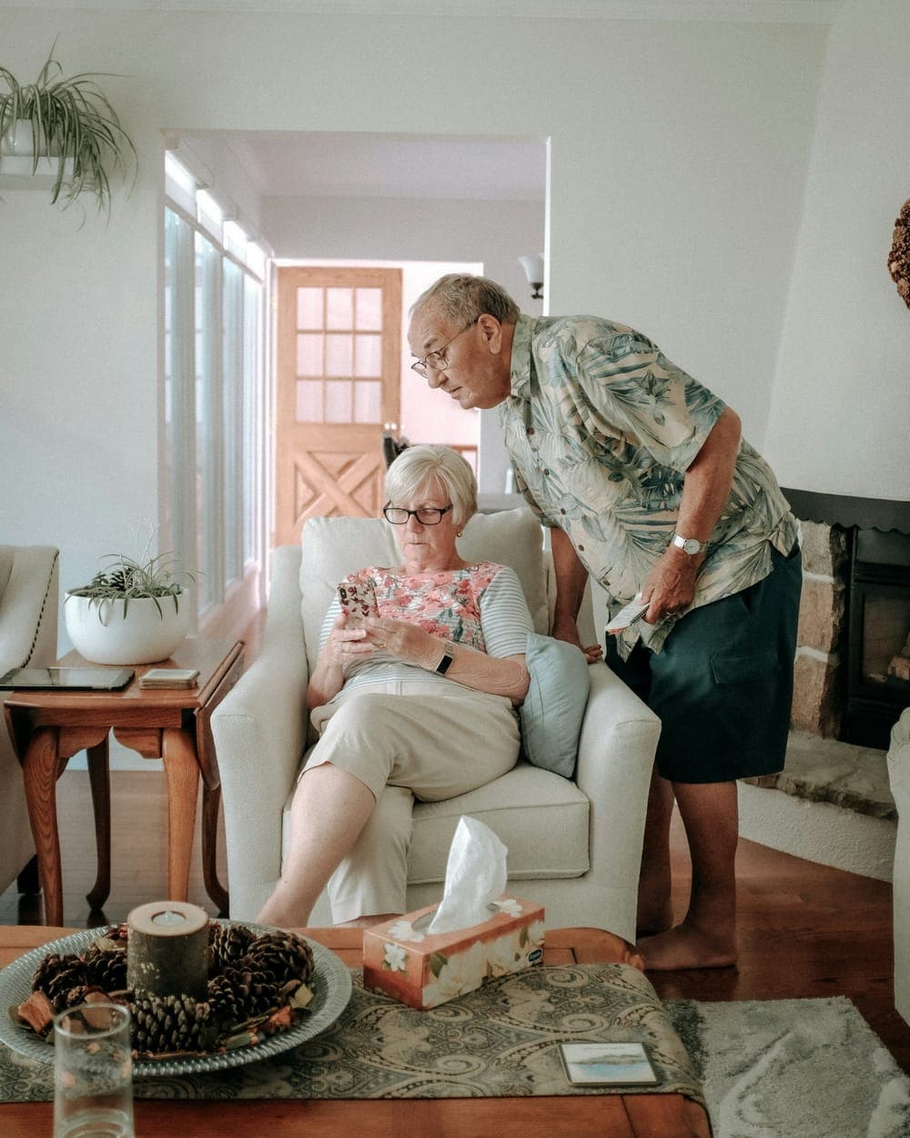 A woman sitting in an armchair in her own front room, with someone leaning in to talk to her.